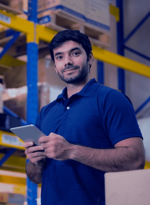 Um homem de camiseta azul segurando um
            tablet,atrás, possuem prateleiras de depósito